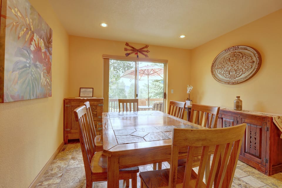A wooden dining table with six chairs sits in a brightly lit room with an ornate wall decoration and a large sliding glass door leading to an outdoor area. Art and furniture adorn the walls and floors.