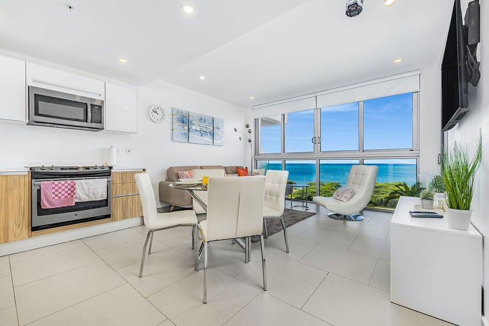 Bright, open living space with sleek white kitchen, cozy dining nook, and sweeping ocean views through floor-to-ceiling windows—your perfect seaside escape.