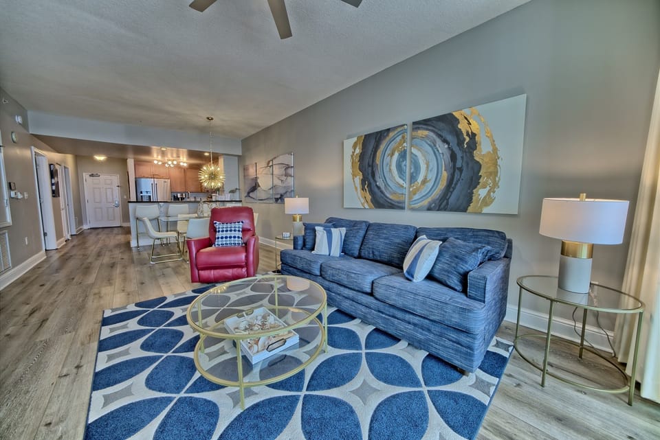 Relax in your stylish open-concept living space featuring a plush blue sofa, contemporary artwork, and seamless flow to your modern kitchen.