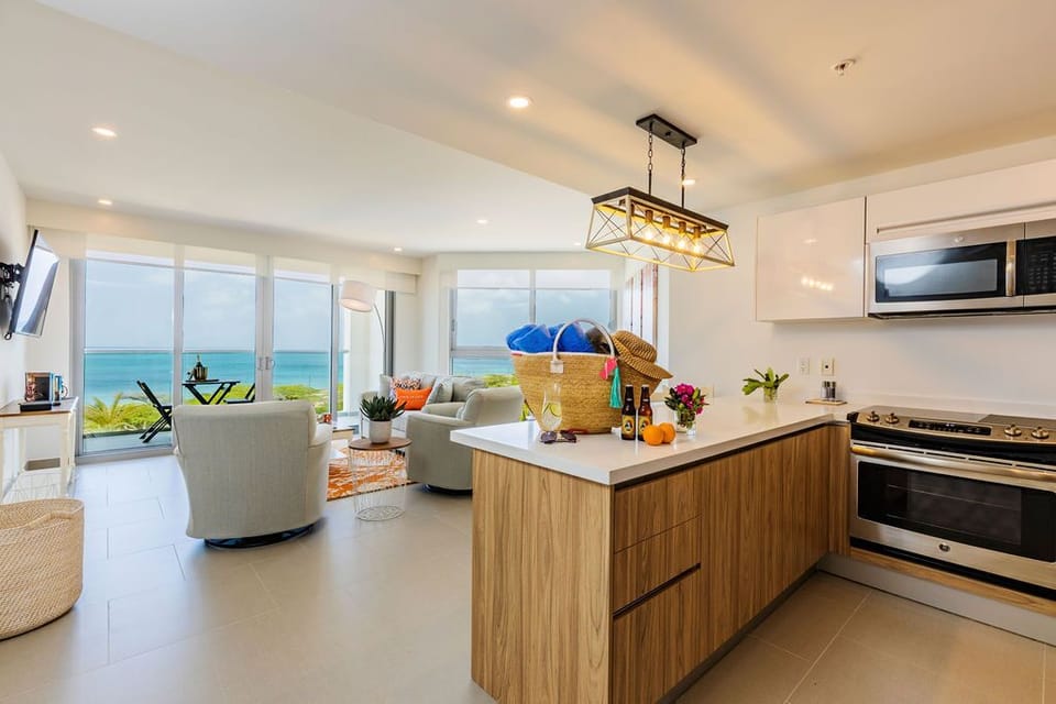 Step into your bright, modern haven where the kitchen flows seamlessly into the living space. Through floor-to-ceiling windows, turquoise waters and coastal greenery frame your days, creating a serene backdrop for cooking, relaxing, and savoring every mome