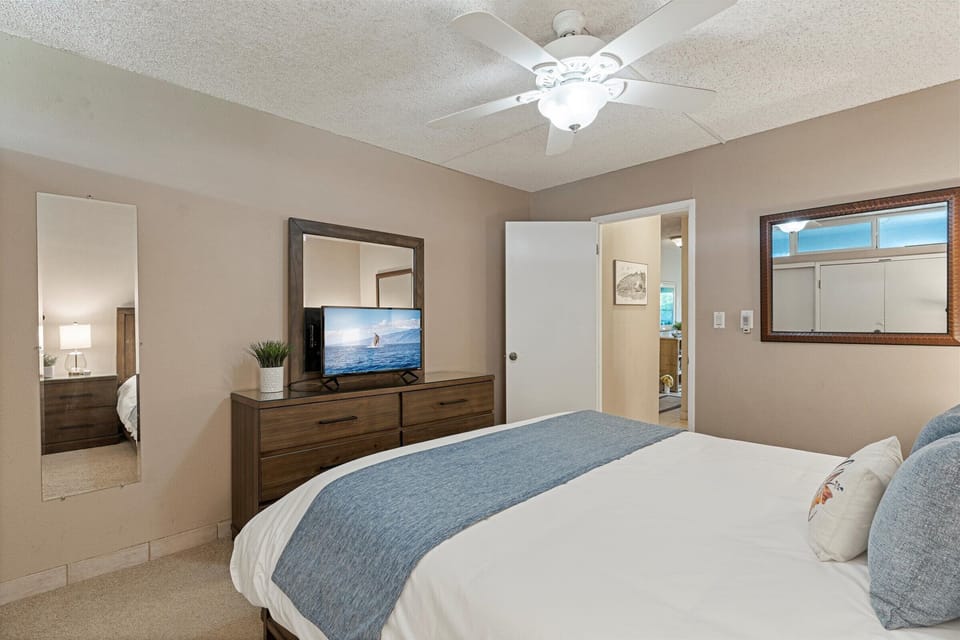 Sink into your cozy bed under the gentle ceiling fan. The spacious bedroom features coastal vibes with ocean scenes on the TV, relaxing neutral tones, and easy access to connecting spaces.