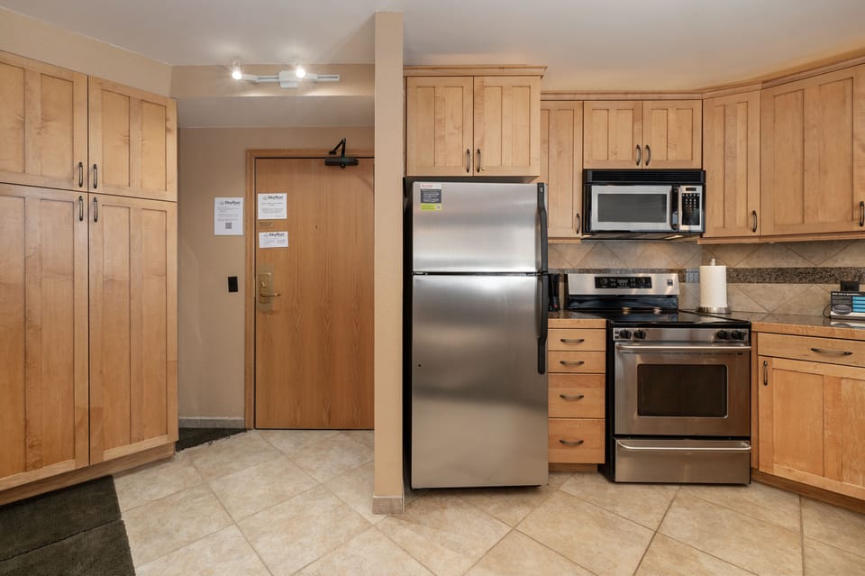 A modern kitchen featuring light wooden cabinets, a stainless steel refrigerator, oven, microwave, and tiled floor. A wooden door with notices attached is visible, along with overhead lighting.