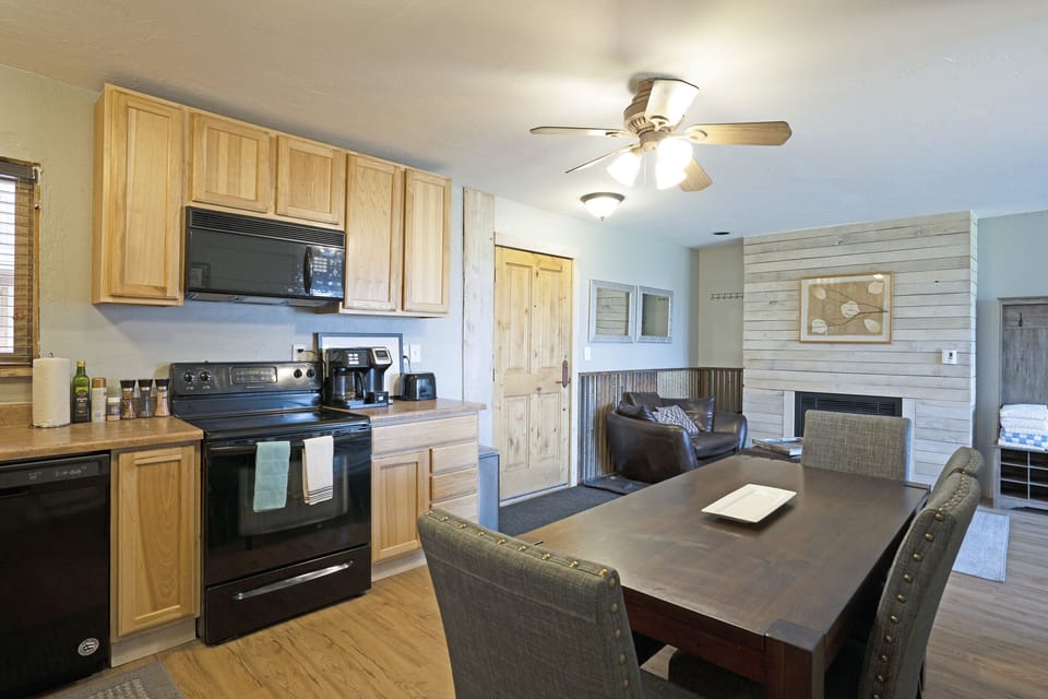 A kitchen and dining area with light wood cabinets, black appliances, and a dark wood dining table. A living room with a fireplace and armchairs is visible in the background.