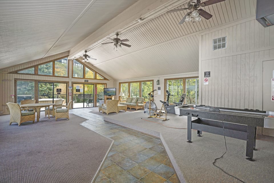 A spacious recreational room with large windows, a foosball table, exercise bike, chairs, and tables. Ceiling fans and overhead lights provide illumination. The room has a mix of carpet and tile flooring.