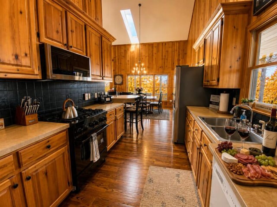 Fully equipped kitchen with natural wood cabinetry and modern appliances flows into the dining area with stunning fall foliage views through large windows.