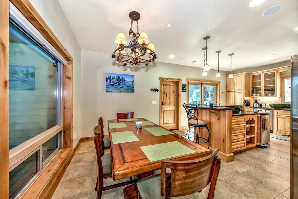 Dining area with a wooden table set for six beneath a metal chandelier, adjacent to a kitchen island with granite counters.