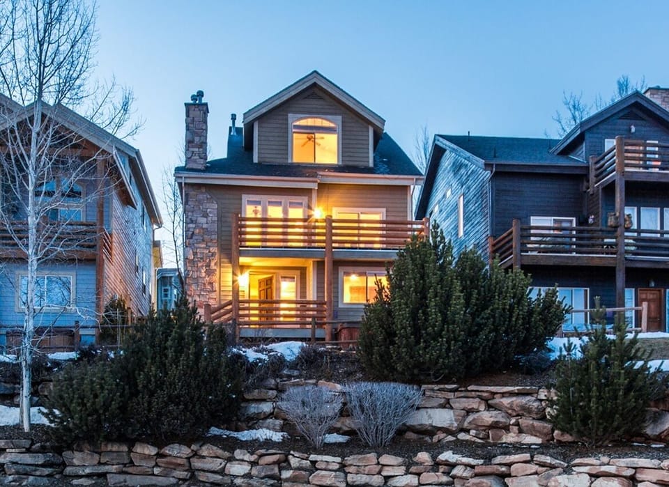 Stunning view of the home's exterior at dusk, showcasing its warm glow and charming architecture. Nestled among tall trees and a rock-lined yard, this beautiful mountain retreat offers comfort, privacy, and inviting curb appeal.