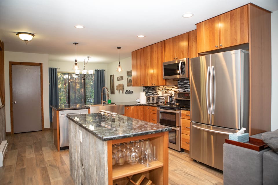 Modern kitchen with wooden cabinets, stainless steel appliances, a marble island with a wine rack, and a backsplash with patterned tiles. There are pendant lights and large windows.