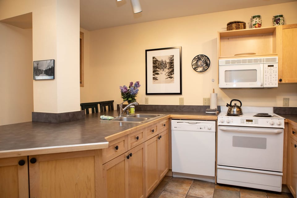 A compact kitchen features wooden cabinetry, a countertop, a sink with a floral arrangement, a dishwasher, a stove, a microwave, and decorative items. A framed picture and wall art decorate the space.