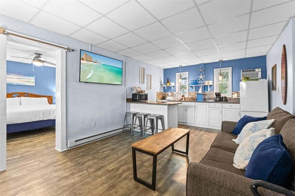Bright living room with smart TV, ceiling fan, cozy sofa seating, and a pass-through bar to the kitchen — a relaxed coastal space to unwind after the beach.