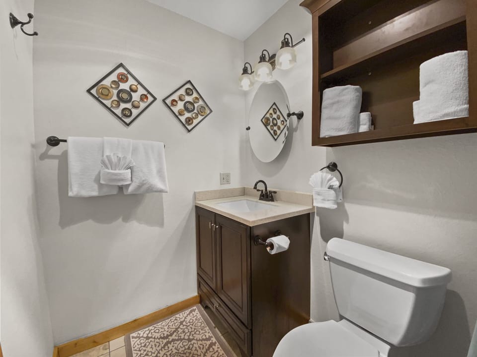 A small bathroom with a sink and mirror, a toilet, towel rack with folded towels, wooden shelves, and wall decorations featuring circular items.