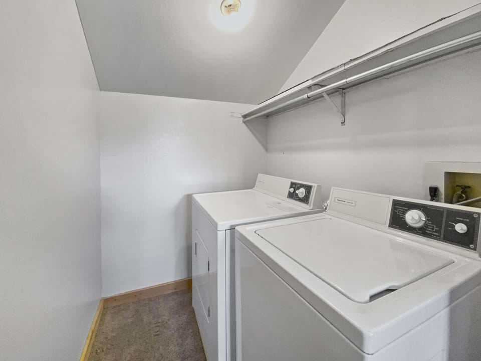 A small laundry room with a white washer and dryer. There is a shelf above the appliances and minimal additional space within the room. Walls and ceiling are white, and the floor is carpeted.
