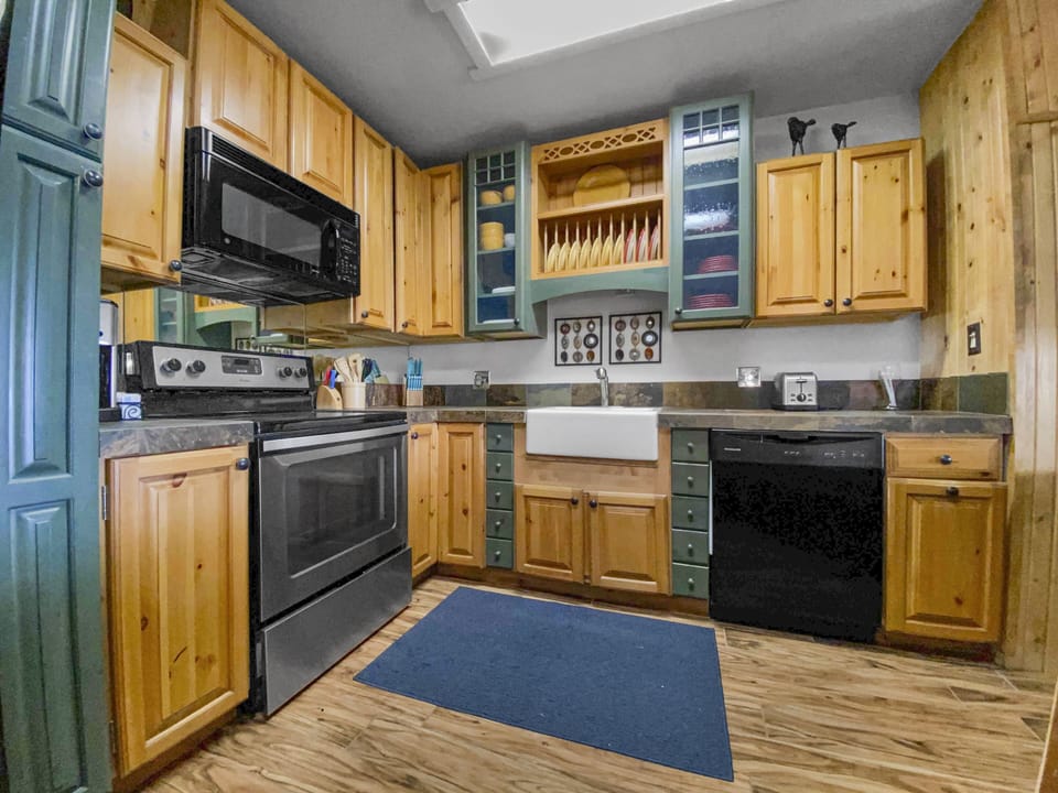 A kitchen with wooden cabinets, black countertops, a stainless steel stove, a black microwave, a white farmhouse sink, a black dishwasher, and a blue mat on a wooden floor.