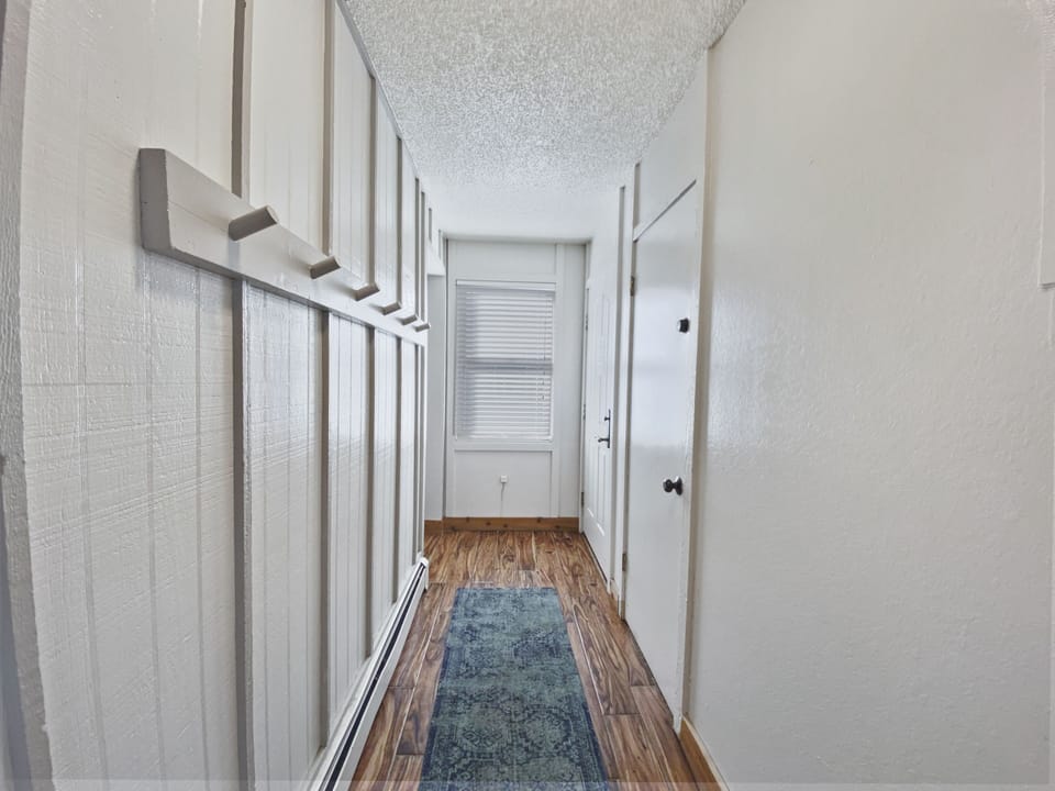 A narrow hallway with white walls, wooden flooring, a blue rug, and a window at the end. Several hooks are mounted on the left wall, and two closed doors are visible on the right.