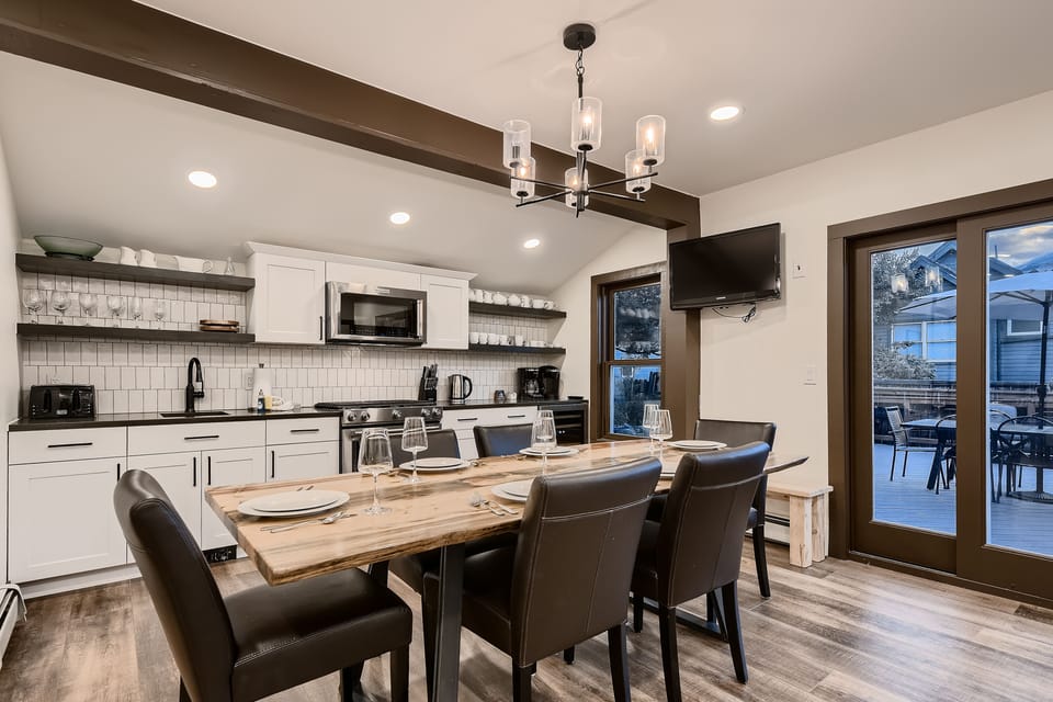 Modern kitchen and dining area with white cabinetry, a wooden dining table set for six, and a wall-mounted TV. Glass doors open to an outdoor seating area.