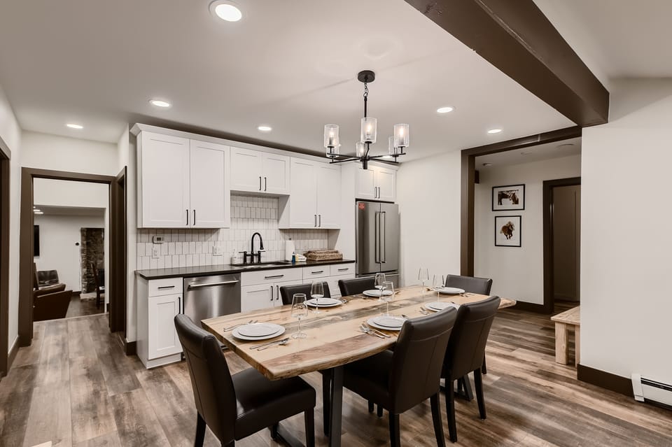 A modern kitchen and dining area with white cabinets, stainless steel appliances, a wooden dining table set for six, and contemporary lighting.