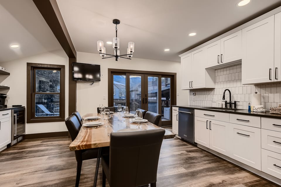 Modern dining room and kitchen area featuring a wooden dining table with six black chairs, white cabinets, stainless steel appliances, a mounted TV, and glass doors opening to an outdoor patio.