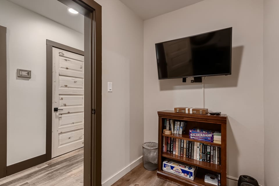 A corner of a room with a mounted flat-screen TV above a small bookshelf containing books and a board game, next to a doorway leading to a hallway with a closed door.