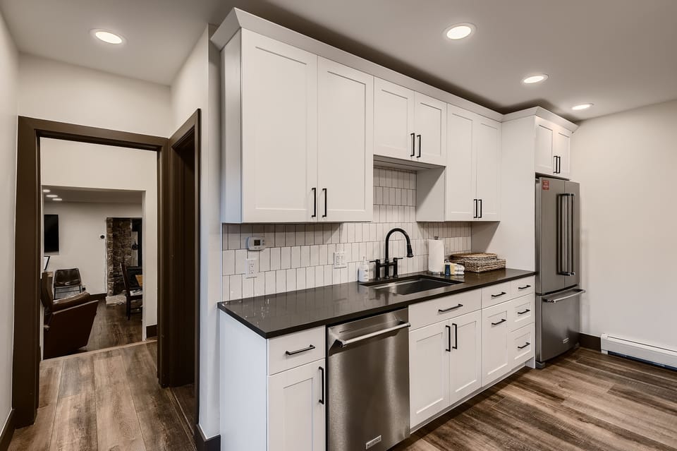 A modern kitchen featuring white cabinets, a black countertop, a stainless steel dishwasher, sink, and refrigerator. The kitchen has a white tiled backsplash and wood flooring. A hallway is visible in the background.