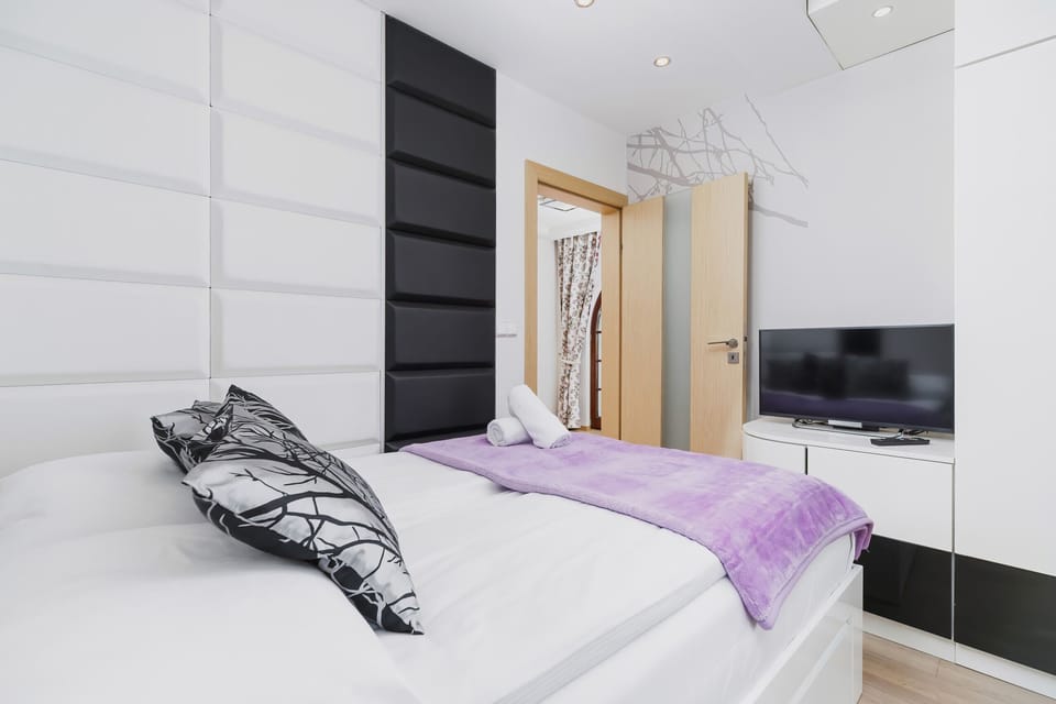 The bedroom features a comfortable bed, soft violet accents, and a flat-screen TV mounted on the wall for evening entertainment.