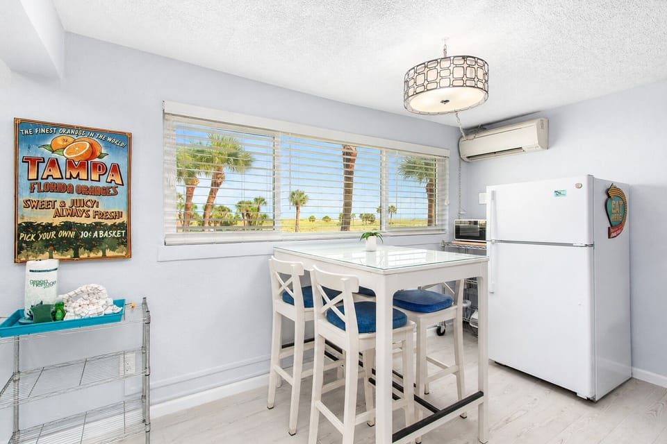 Cozy dining nook with a glass-top table seating four, bright window views, and a full-size fridge nearby. The perfect spot to enjoy meals together after a day at the beach.