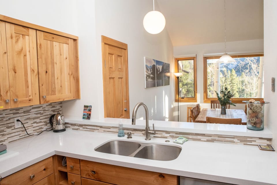 A kitchen with wooden cabinets, a double sink, and a countertop looking into a dining area with a window, showing a scenic outdoor view. The wall features a scenic photograph and hanging light fixtures.