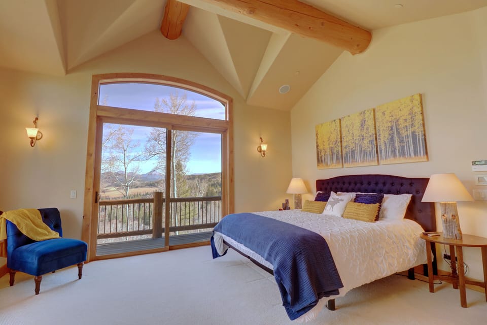 A bedroom with a large window, king-sized bed, blue chair, nightstands, lamps, and wall art depicting trees. The window offers a view of an outdoor landscape.