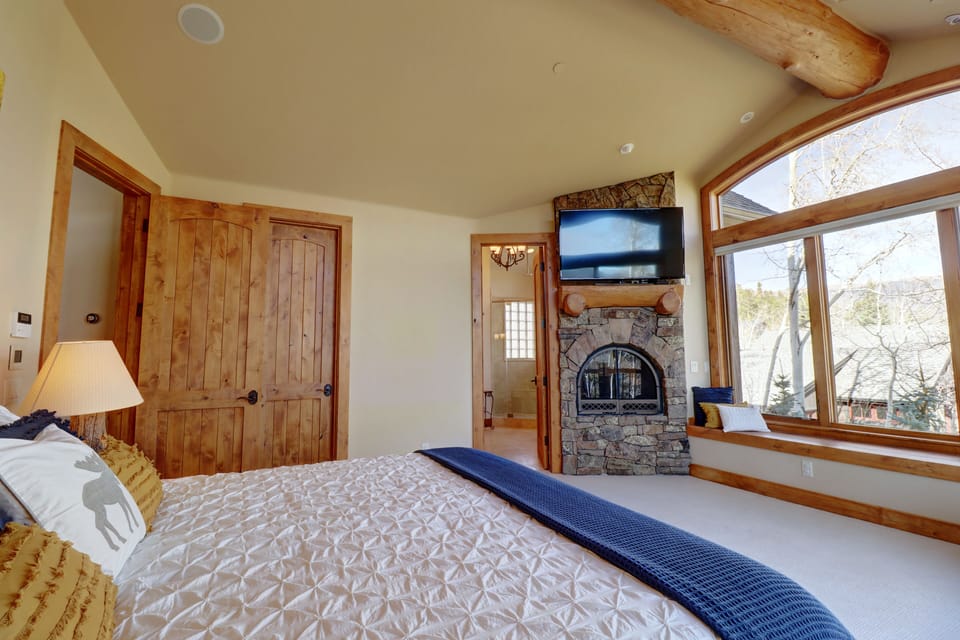 A bedroom with a large bed, wooden doors, a stone fireplace with a mounted TV, and a large window offering a view of trees and a mountainous landscape.