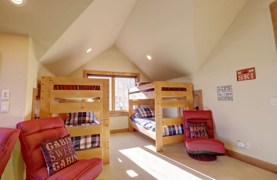 A cozy loft room with two wooden bunk beds, plaid bedding, two red chairs, and window allowing natural light. Text signs on the walls read "Cabin Sweet Cabin" and "Ski.