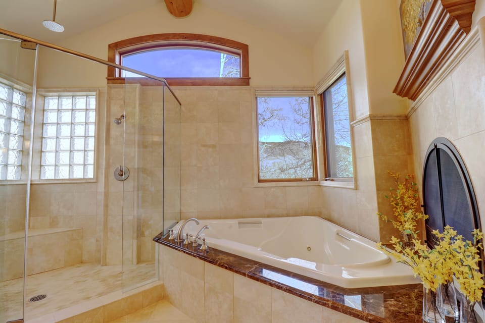A bathroom with a large corner bathtub, glass-enclosed shower, and windows providing natural light. Decor includes a plant on the bathtub's edge. The space has beige tiles and wood trim.