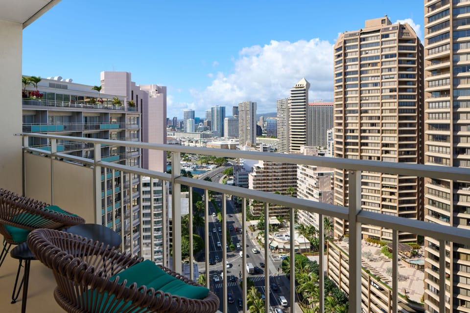Elevated balcony showcasing a sweeping cityscape with high-rise buildings, lush palms below, and a vibrant urban skyline stretching toward distant mountains under bright blue skies.