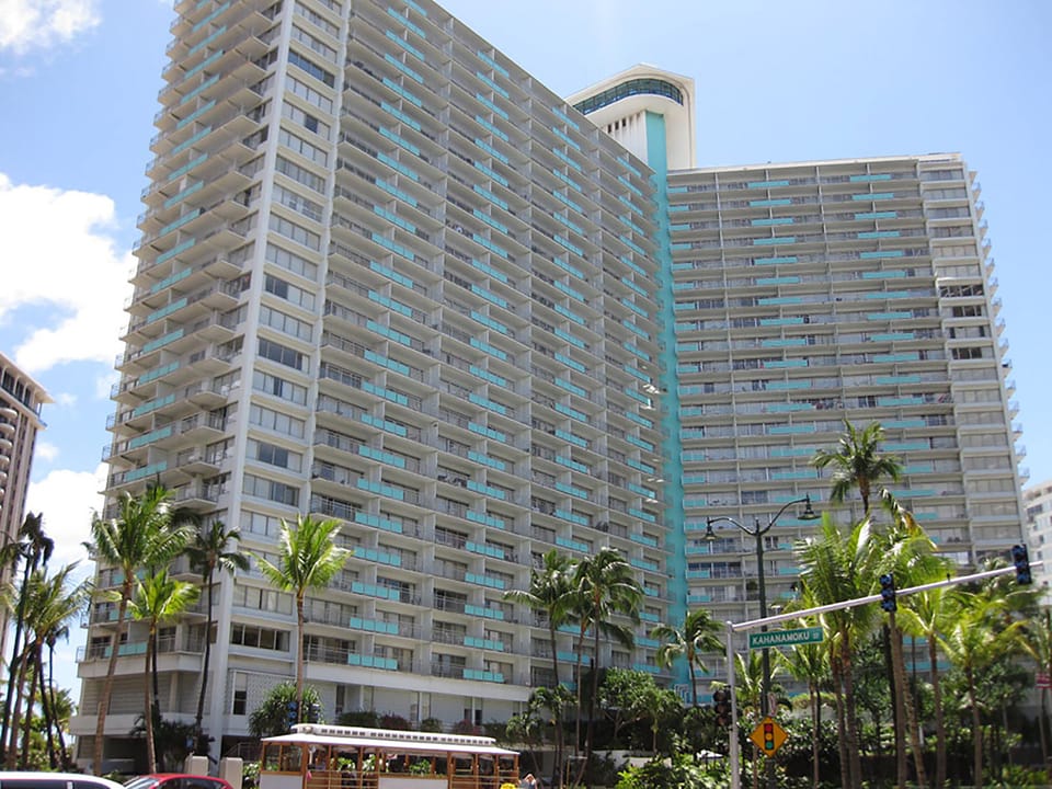 This high-rise beachfront building features numerous balconies with stunning ocean views, surrounded by swaying palms and located just steps from Waikiki's sandy shores and vibrant neighborhood.
