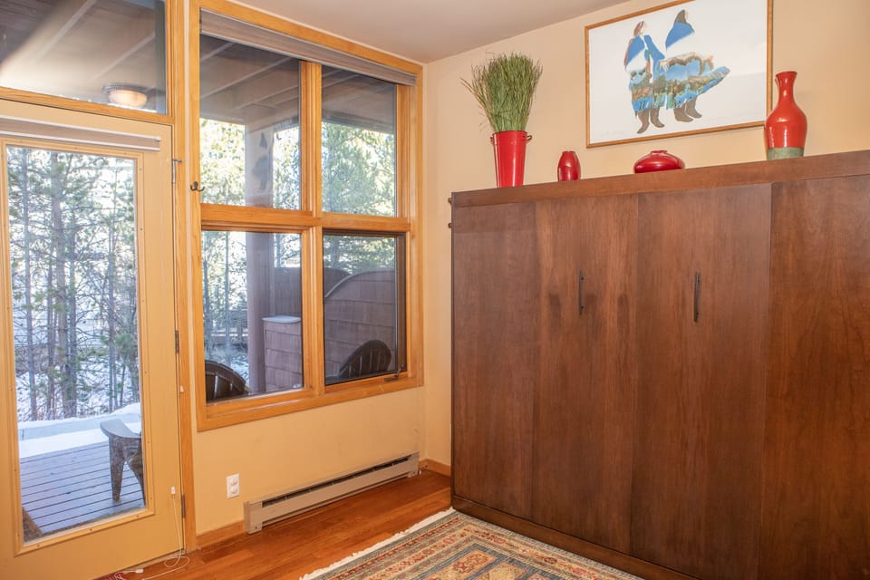 A room with a large wooden cabinet, a window with forest view, colorful rug, red vases on the cabinet, and a framed artwork on the wall.