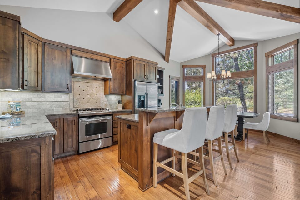 Gather at the heart of this stunning chef's kitchen, where vaulted ceilings with exposed wood beams meet modern appliances and a welcoming breakfast bar, all bathed in natural light from the surrounding trees.