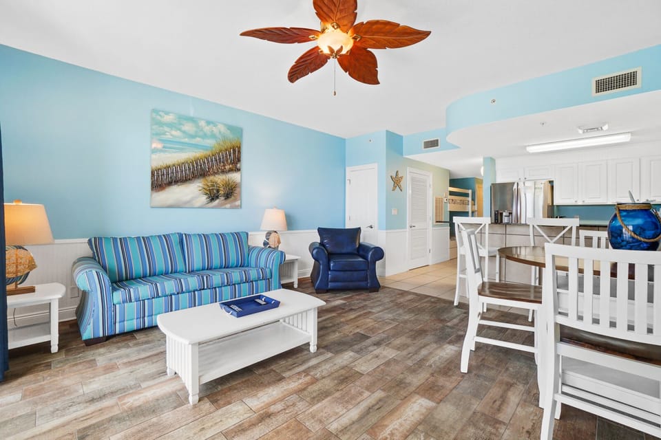 Relax in your bright, coastal-themed living space featuring comfy striped seating, tropical ceiling fan, and open layout connecting to your fully equipped kitchen.