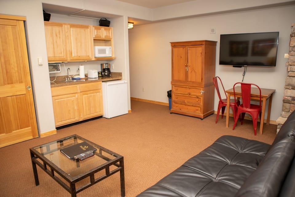 A small living area with a kitchenette, featuring wooden cabinets, a mini-fridge, a microwave, a coffee maker, a television on the wall, a coffee table, a sofa, and a wooden armoire.