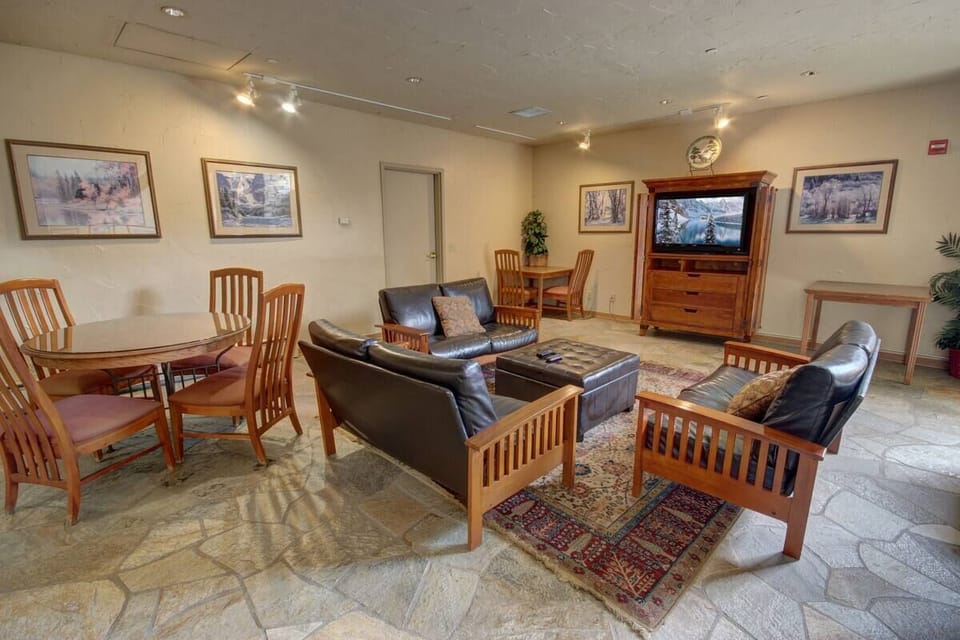 A living room with stone flooring, wooden furniture, leather seats, a wooden dining table with chairs, framed artworks on the walls, and a wooden TV cabinet with a clock on top.
