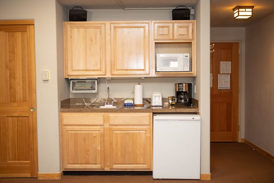A compact kitchenette with wooden cabinets, a small sink, microwave, toaster oven, coffee maker, and mini-fridge. A door is visible to the right, and lighting fixtures are attached to the ceiling.