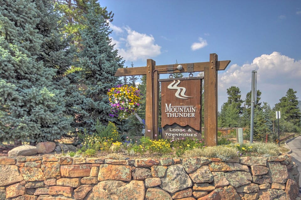 A wooden sign for "Mountain Thunder Lodge & Townhomes" is mounted on stone, surrounded by pine trees and flowers. A road is visible in the background.