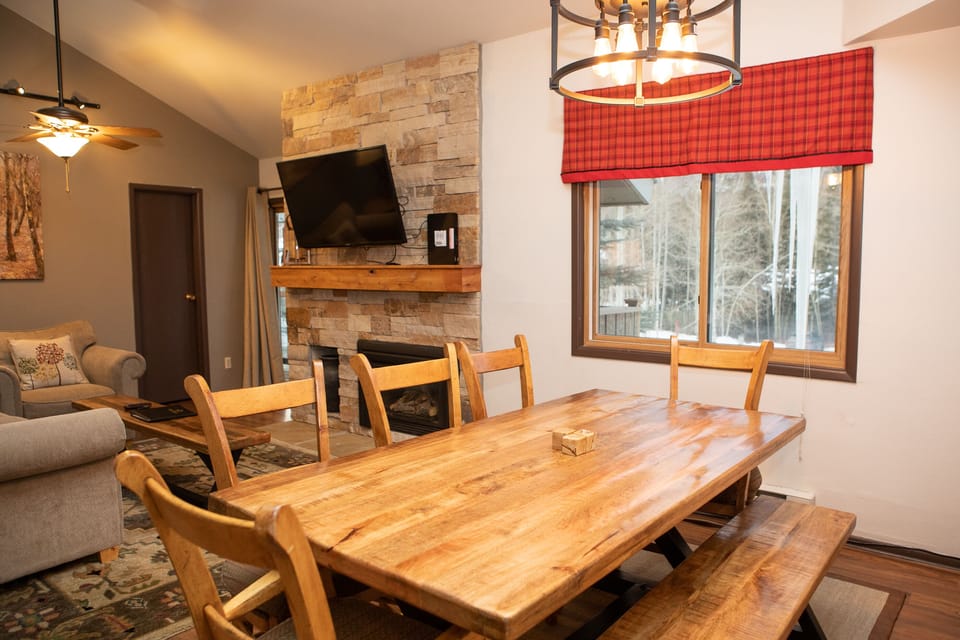 A living room with a wooden dining table and bench, stone fireplace, wall-mounted TV, and a window with a red plaid curtain, overlooking a snowy outdoor view.