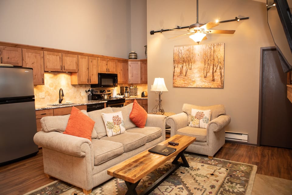 Living room and kitchen with wood cabinets, a fridge, stove, and counter. Two beige sofas with orange accent pillows face a wooden coffee table. A ceiling fan and framed artwork are on the wall.