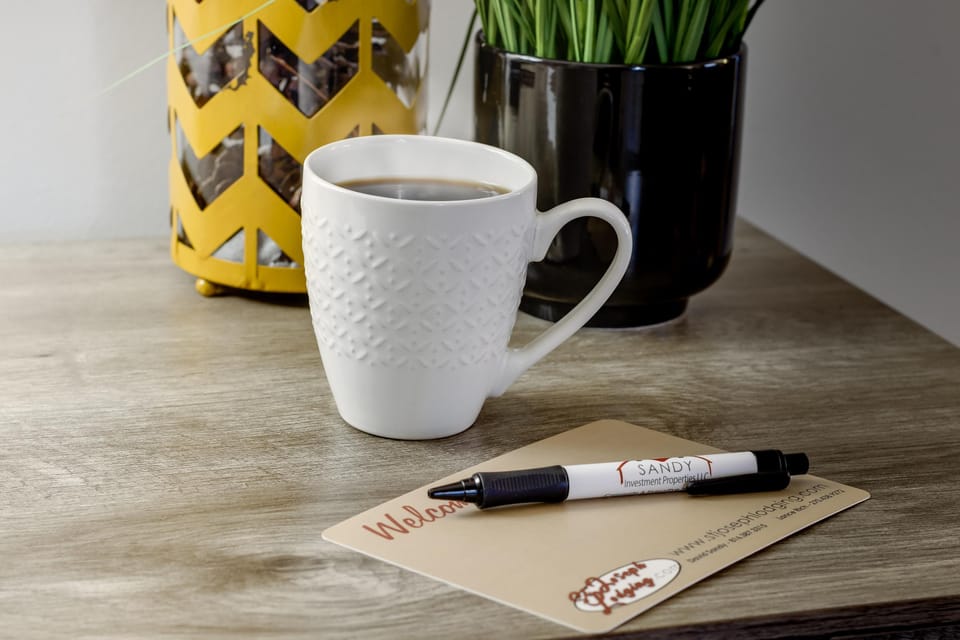 Close-up of a white textured ceramic mug filled with coffee on a wood-grain tabletop beside a pen and welcome card.