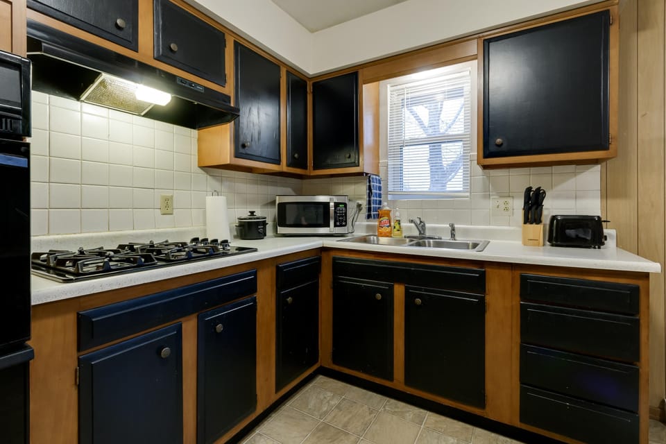 L-shaped kitchen with black and wood cabinetry, white tile backsplash, and a double stainless steel sink beneath a window.