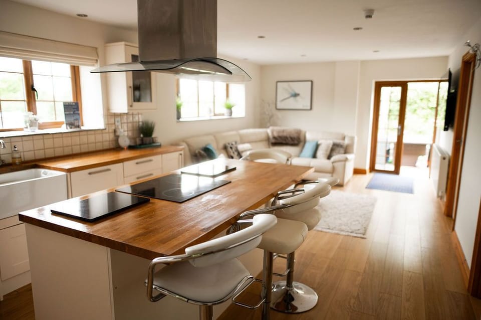 A modern, open-concept kitchen and living area featuring a sleek kitchen island with a wooden countertop and built-in stove, accompanied by contemporary bar stools. The space is illuminated by natural light streaming in through large windows and a glass door, leading to an outdoor area. The adjacent cozy living space includes a cream-colored sectional sofa adorned with soft cushions, creating an inviting atmosphere.
