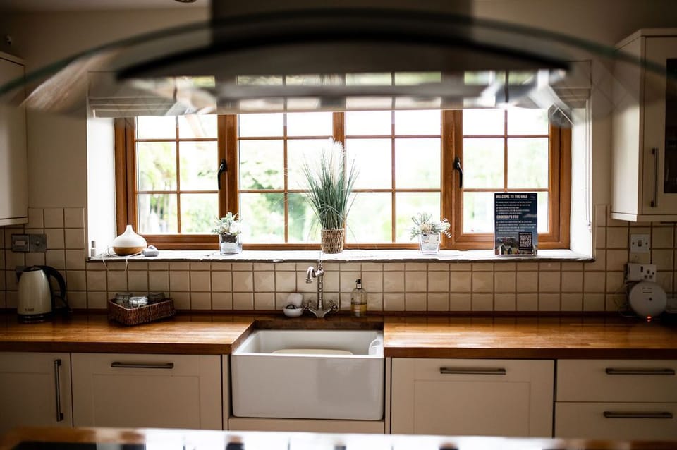 A charming kitchen sink area, featuring a large farmhouse-style sink set against a backdrop of beige tiled walls. The wooden countertop provides warmth and character, while a series of potted plants on the windowsill add a touch of greenery and freshness. Natural light streams in through the large, divided-light windows, illuminating the space. A few kitchen essentials, including a kettle and a woven basket, are neatly arranged on the counter, maintaining a clean and organized look. This cozy and inviting setup exudes a homely and functional atmosphere.