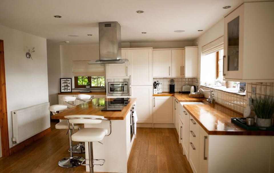 A bright and contemporary kitchen featuring cream cabinetry and sleek wooden countertops. The central island doubles as a breakfast bar with modern bar stools and a built-in stovetop, complemented by a stylish range hood. The kitchen is equipped with stainless steel appliances, including a microwave and a toaster, and boasts ample counter space for meal preparation. Large windows allow plenty of natural light to flood the room, enhancing its warm and inviting ambiance.