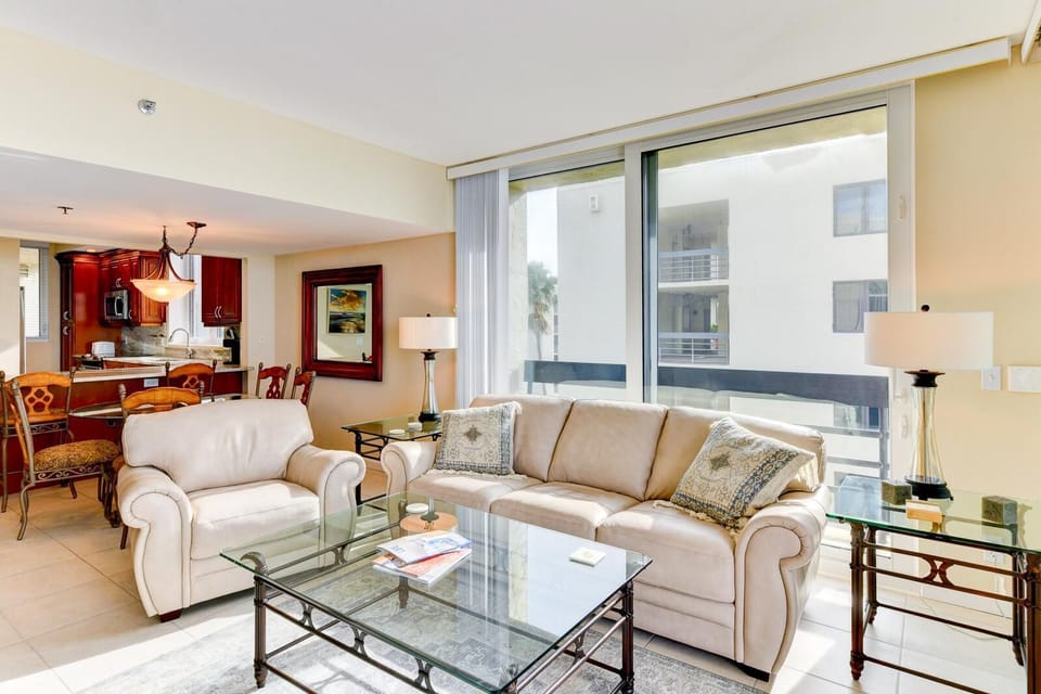 Natural light fills the living room with beige leather seating, glass-topped tables, and a view of the balcony sliding doors.