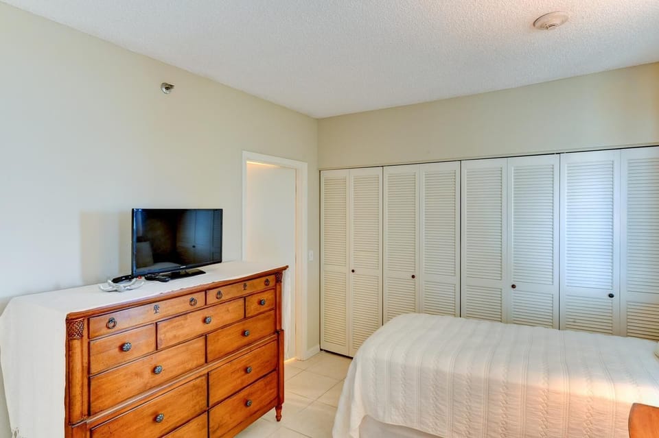 Flat-screen TV rests on a carved wooden dresser next to white louvered closet doors and beige tiled flooring.