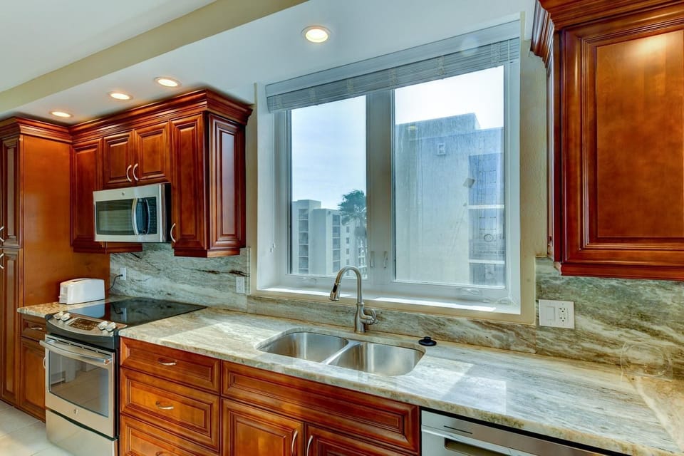 Kitchen features polished granite countertops, stainless steel appliances, and warm wood cabinetry beneath a large window.