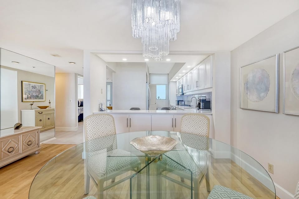 Glass-top dining table with patterned chairs beneath a tiered glass chandelier and light hardwood floors.